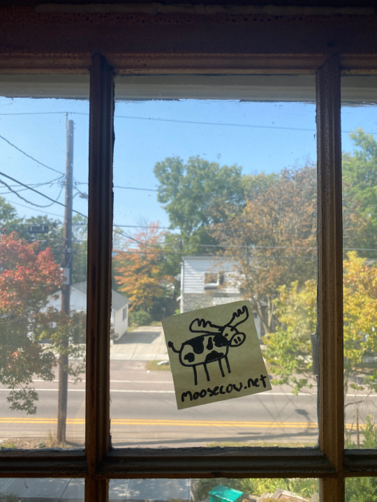 A crude sharpie on post-it-note drawing of a cow with antlers.  The post-it-note is on a window, with an early fall street scene behind it